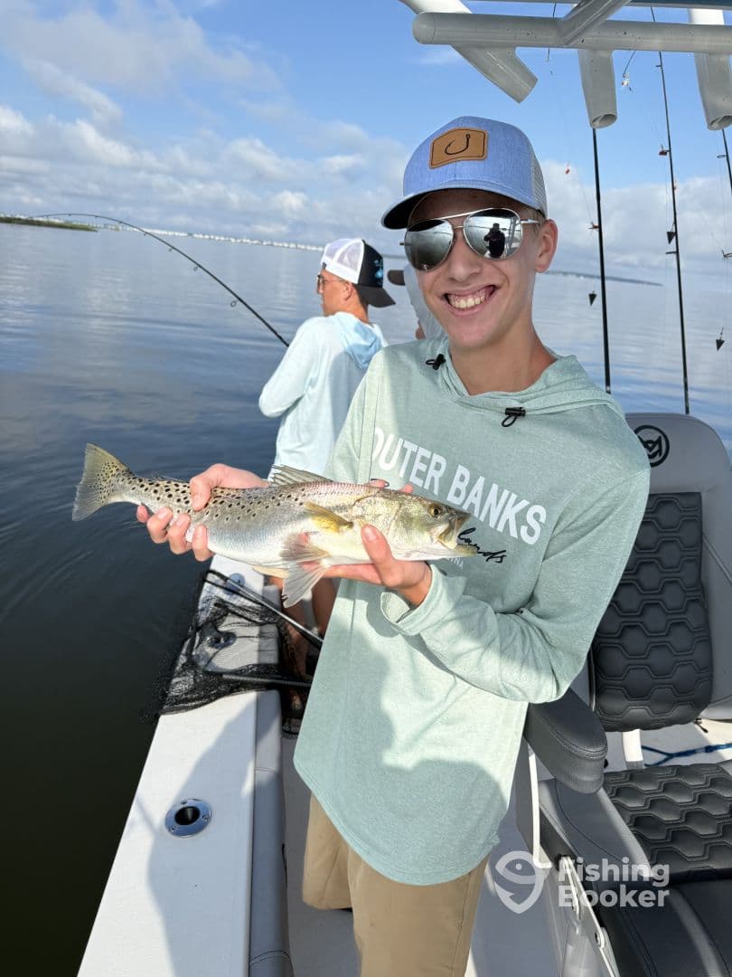 A young angler proudly displaying a Speckled Trout while fishing from a boat, enjoying a sunny day on the water.