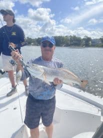 An angler proudly displaying a large Redfish while fishing on a sunny day, with another fisherman in the background.