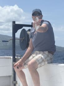 An angler proudly displaying a small fish while seated on a boat, with a scenic ocean backdrop.