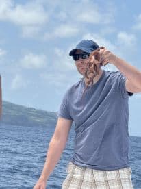 Angler proudly displaying a freshly caught octopus while fishing off the coast, with a scenic backdrop of green hills and blue waters.