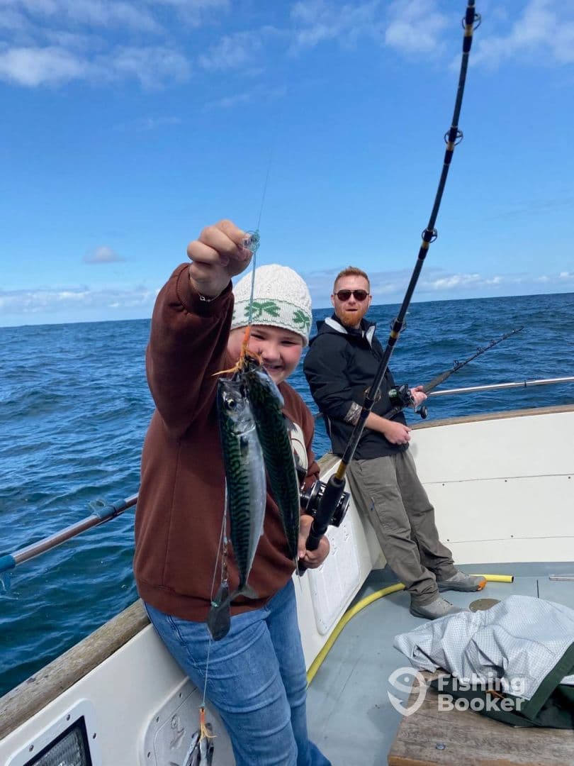 A young angler proudly displaying two Mackerel while fishing on a boat, with a scenic ocean backdrop.