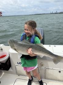 A young angler joyfully holding a large Striped Bass while fishing on a boat, showcasing a successful day on the water.