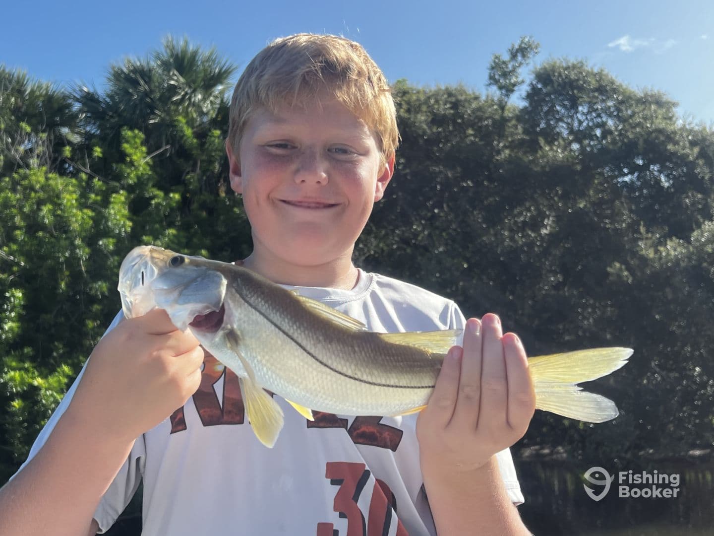 A young angler proudly displaying a Snook caught during a sunny day fishing trip, surrounded by lush greenery.