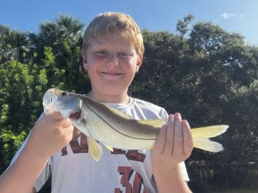 A young angler proudly displaying a Snook caught during a sunny day fishing trip, surrounded by lush greenery.
