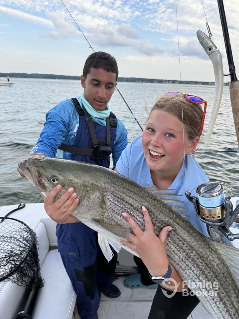 Two young anglers proudly displaying a large Striped Bass while fishing on a boat, showcasing a fun day on the water.