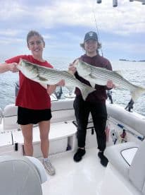 Two anglers proudly displaying their catch of striped bass while aboard a fishing boat, enjoying a day on the water.