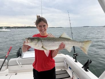 A young woman proudly displaying a large Striped Bass while fishing on a boat, showcasing a successful day on the water.