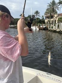 A young angler reeling in a fish while fishing in a scenic waterway, surrounded by palm trees and upscale homes.