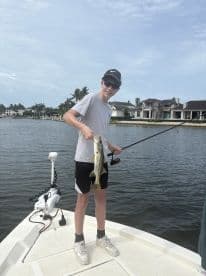 A young angler proudly displaying a Snook while fishing from a boat in a scenic waterfront setting.