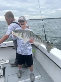 A young angler proudly displaying a large Striped Bass while fishing on a boat, showcasing a fun day on the water.