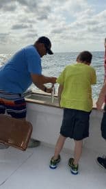 A young boy and an adult are engaged in fishing, with the adult helping the boy handle a fish on a boat, showcasing a hands-on learning experience at sea.