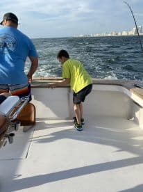 A young boy leans over the side of a boat, observing the water, while an adult stands nearby. The backdrop features a coastal city skyline under a clear sky.
