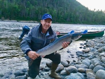 An angler proudly displaying a large Atlantic Salmon while kneeling on the rocky shore of a river, with a canoe visible in the background.