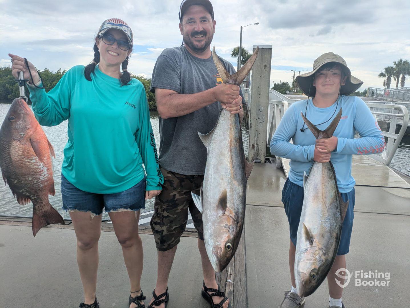 Family proudly displaying their catch, including a large Snapper and Amberjack, at the dock after a successful fishing trip.