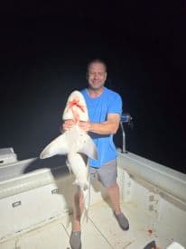 Angler proudly displaying a large shark on a boat during a night fishing trip.