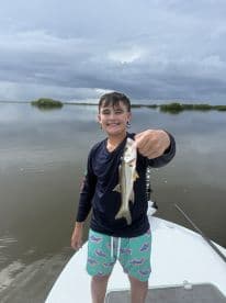 A young angler proudly displaying a small Snook while fishing on a calm waterway, surrounded by mangroves.