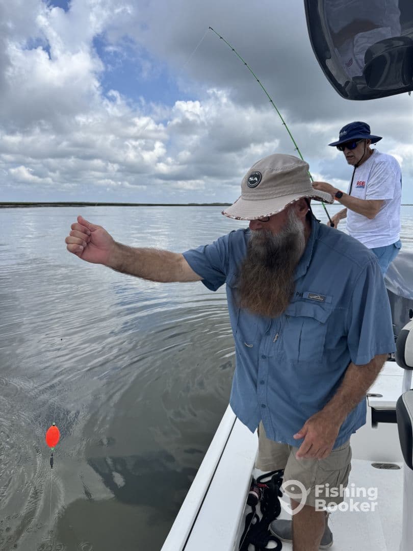 Two anglers actively fishing on a boat, with one reeling in a line while the other assists. The scene captures the excitement of fishing in a calm water environment.