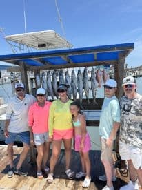 A family poses proudly in front of a catch board displaying several fish, including King Mackerel, after a successful fishing trip.