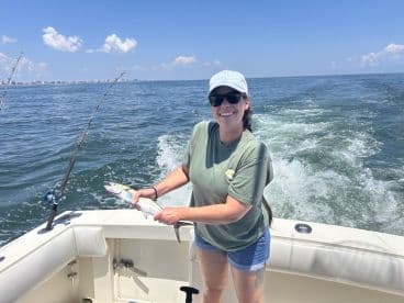 A smiling angler holding a freshly caught fish while enjoying a sunny day on the water, with a coastal city skyline in the background.