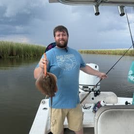 An angler proudly displaying a caught stingray while fishing from a boat in a calm waterway, surrounded by lush greenery.