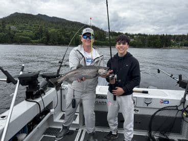 Two anglers proudly displaying a large Salmon on a fishing boat in a scenic coastal environment.
