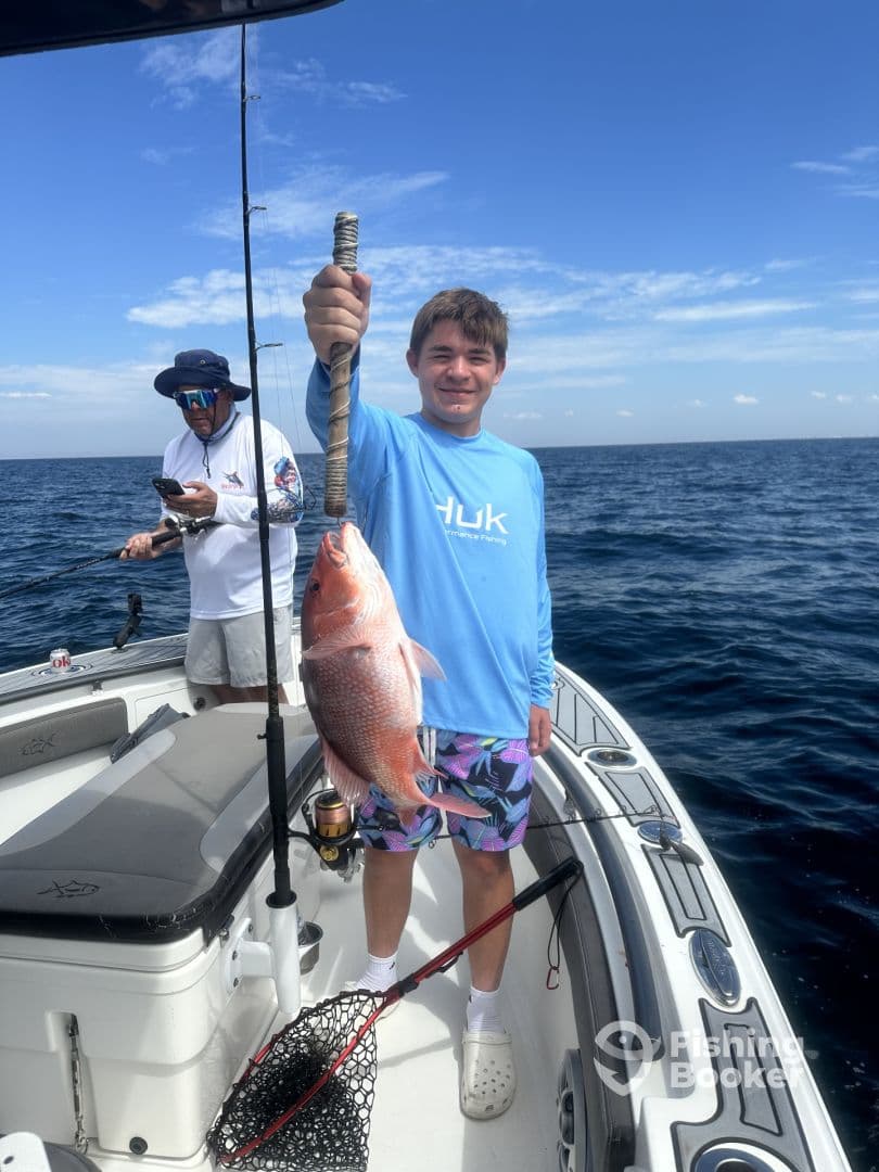 A young angler proudly displaying a large Red Snapper while fishing on a boat, with another angler in the background.