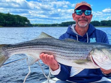 An angler proudly displaying a large striped bass while fishing on a scenic lake, showcasing a beautiful day outdoors.