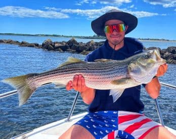 Angler proudly displaying a large Striped Bass while on a fishing trip, with a scenic waterfront backdrop.