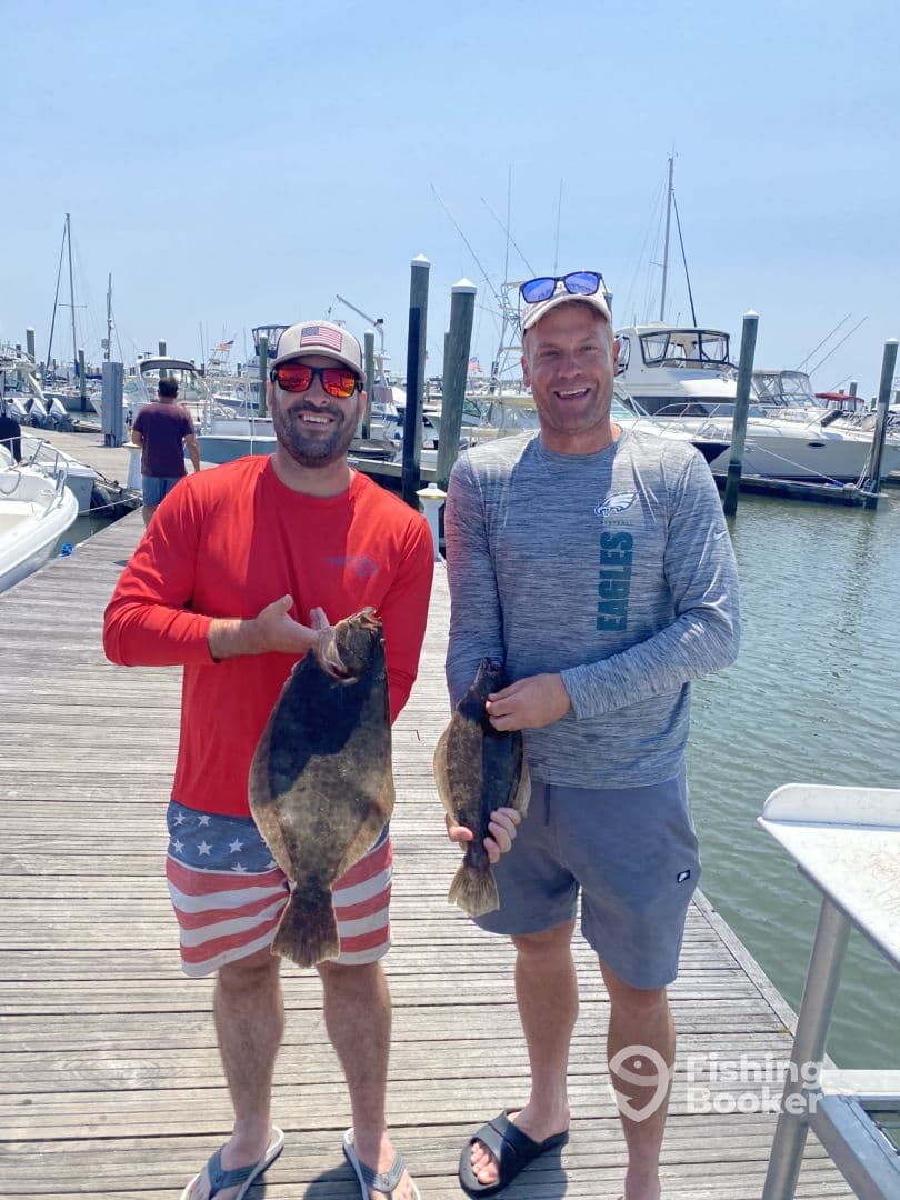 Two anglers proudly displaying their catch of Flounder at a marina, showcasing a successful day of fishing.