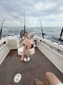 A young girl enjoying her time on a fishing boat, giving a thumbs up while seated in the helm area, with fishing rods visible in the background.