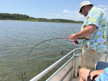 An angler reeling in a catch while fishing from a pontoon boat on a serene lake, surrounded by lush greenery.