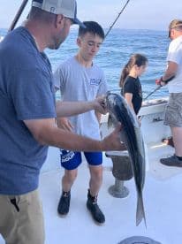 A young angler is holding a large striped bass while fishing on a boat, surrounded by fellow anglers in a coastal environment.