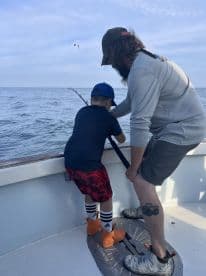 A young angler and an adult are working together to reel in a fish while fishing on a boat, showcasing a bonding moment on the water.