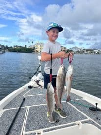 A young angler proudly displaying three Redfish on a boat in a scenic waterfront setting.