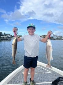A young angler proudly displaying two Redfish while standing on a boat in a scenic waterway.