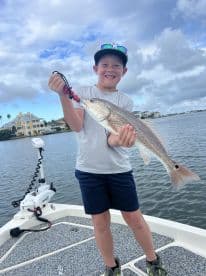 A young angler proudly displaying a Redfish while fishing from a boat in a scenic coastal environment.