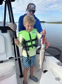 A young boy in a life jacket proudly holding a caught fish while on a boat, enjoying a family fishing trip.
