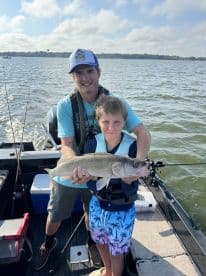 A young boy proudly displays a Walleye he caught while fishing with an adult on a boat, showcasing a fun day on the water.