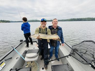 Two anglers proudly displaying a Northern Pike while fishing on a calm lake, with a child in the background casting a line.
