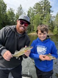 A proud adult and child display their catches of Walleye while fishing on a serene lake surrounded by lush greenery.