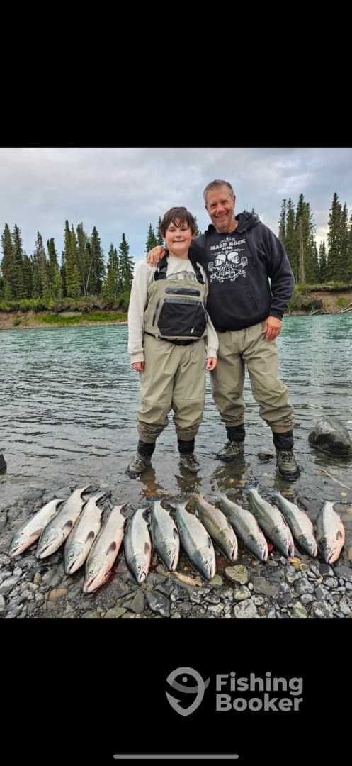 A young angler and an adult proudly display their catch of multiple salmon along a riverbank, showcasing a successful fishing trip.