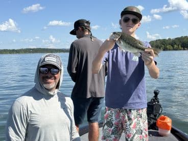 A young angler proudly displaying a large Largemouth Bass while fishing on a scenic lake, with a smiling adult in the background.