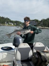 A young angler proudly displaying a striped bass while aboard a fishing boat in a scenic coastal setting.