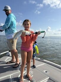A young girl proudly displaying a Speckled Trout while fishing on a boat, accompanied by an adult and another child in a sunny coastal environment.