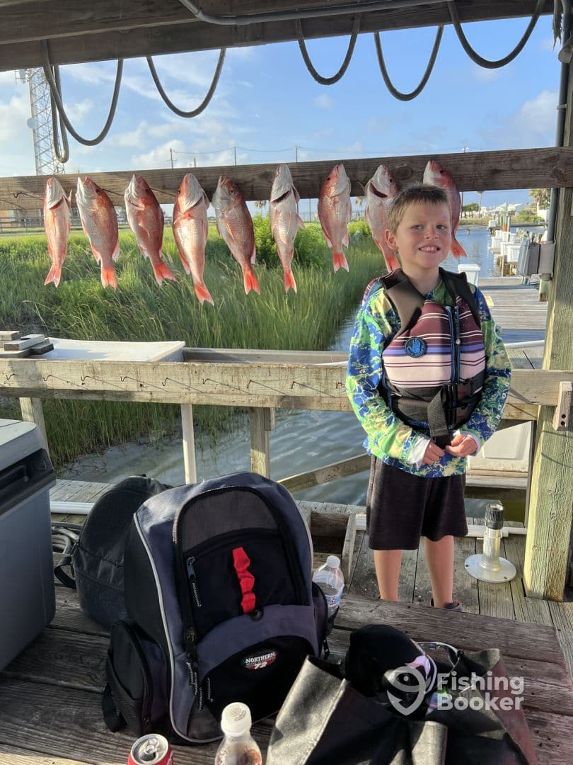 A young boy stands proudly next to a catch of Red Snapper displayed on a board at the dock, showcasing a successful fishing trip.