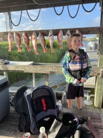 A young boy stands proudly next to a catch of Red Snapper displayed on a board at the dock, showcasing a successful fishing trip.