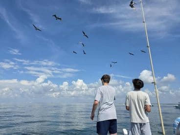 Two young anglers enjoying a fishing trip, observing birds in the sky while waiting for a catch on a calm day at sea.