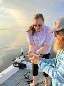 A woman joyfully holds a freshly caught trout while fishing on a calm morning, showcasing a successful catch during a charter trip.