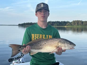 A young angler proudly displaying a large Redfish while fishing on a calm waterway.