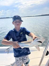 Young angler proudly holding a small shark while on a boat, showcasing a successful fishing trip.